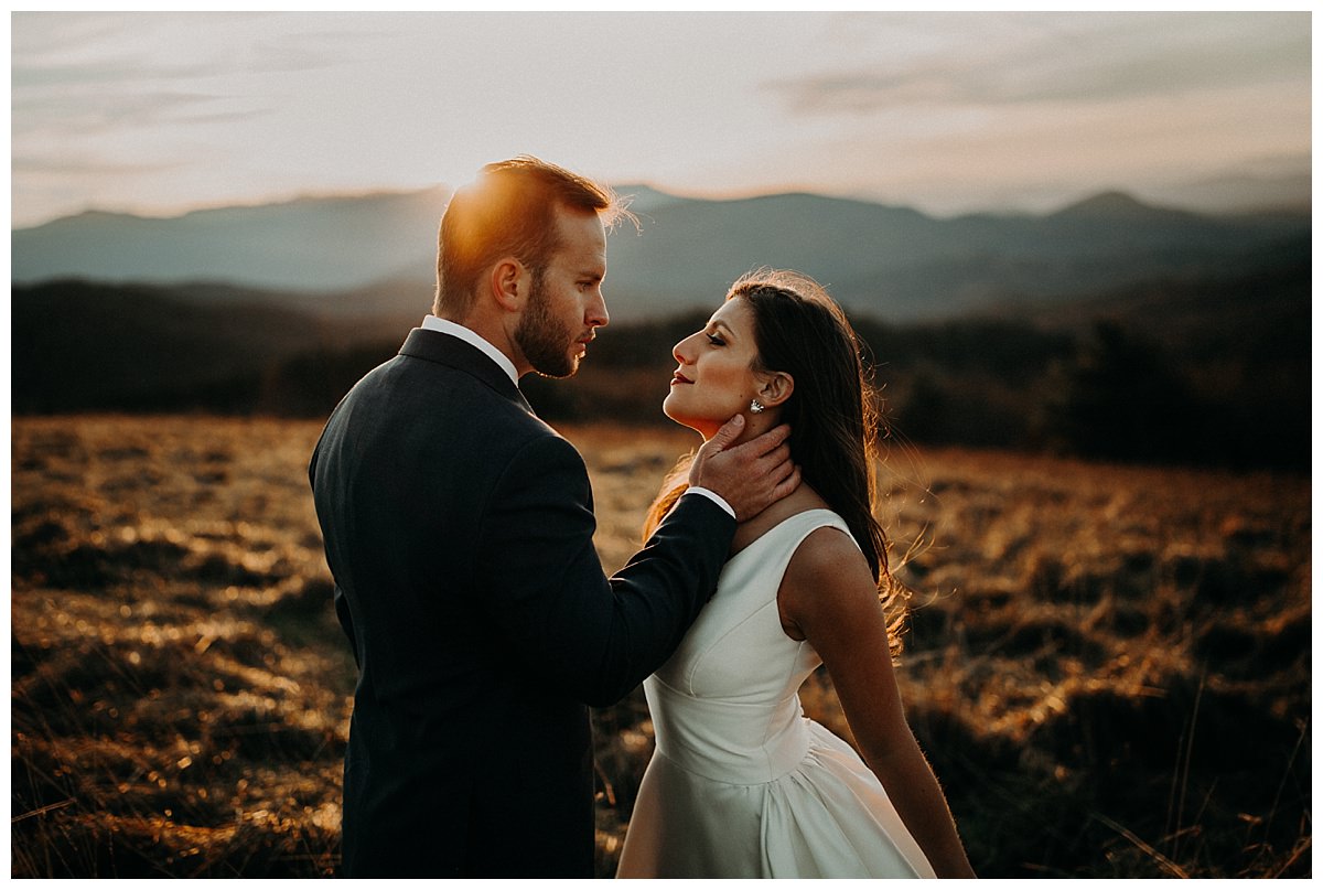 A man in a suit caressing a woman in a beautiful wedding dress at Max Patch overlooking the mountains at sunset.  This elopement photo captures the intimacy between the bride and groom. 