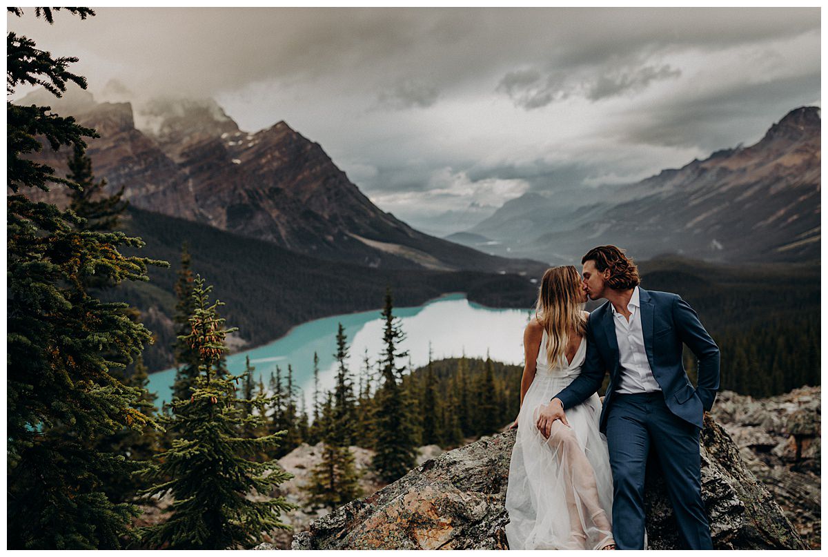 An elopement at Peyto Lake in Banff National Park in Canada.  The bride and groom are sitting on a rock overlooking Peyto Lake and kissing each other.  An alternative to planning your wedding during coronavirus is to plan an intimate wedding or elopement instead. 