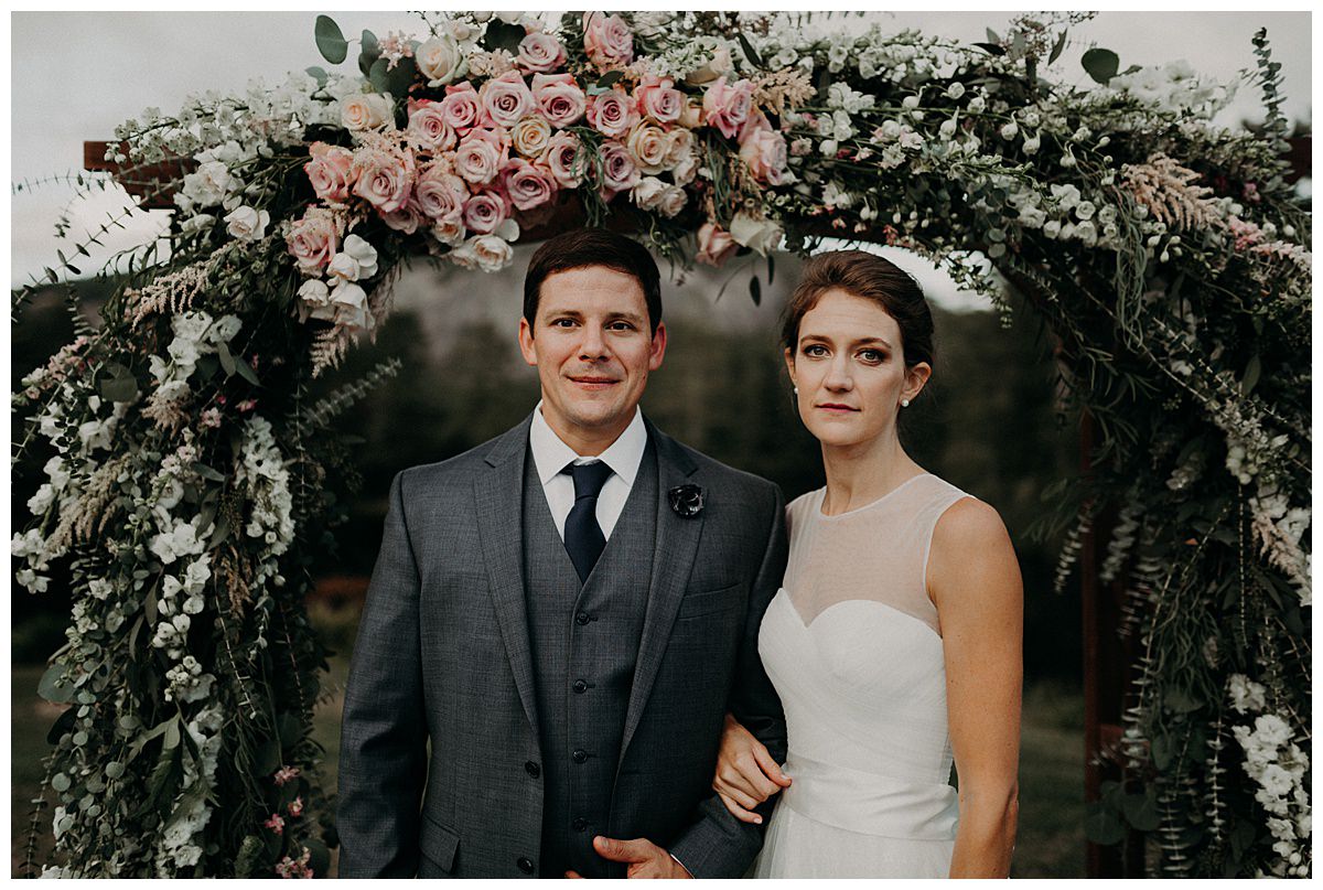 A man and woman in wedding attire are standing below a beautiful flower arbor.  They planned their wedding during coronavirus.  Both are looking pensively at the camera. The photo is edited with a darker, moodier look to it.  