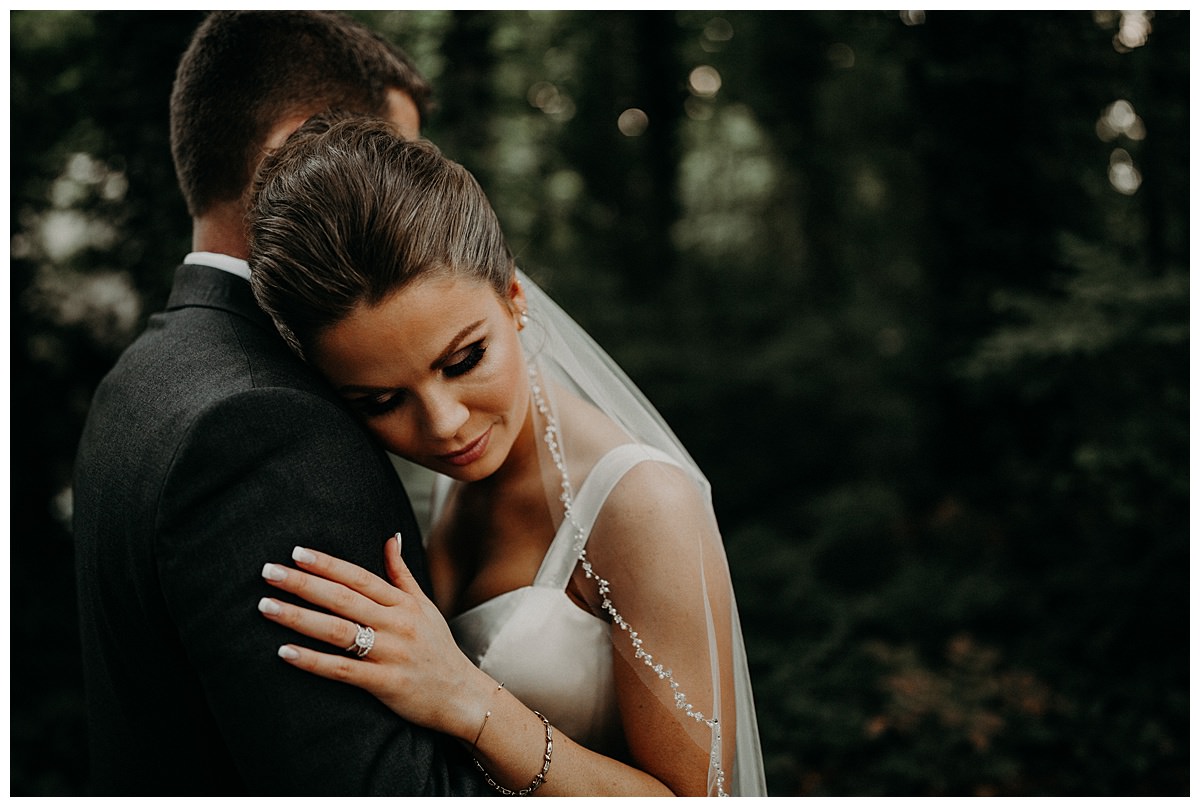 A bride rests on her grooms shoulder with her left hand grabbing his tricep area showing off a magnificent ring.  These two set proper expectations about their wedding during Covid-19.   
