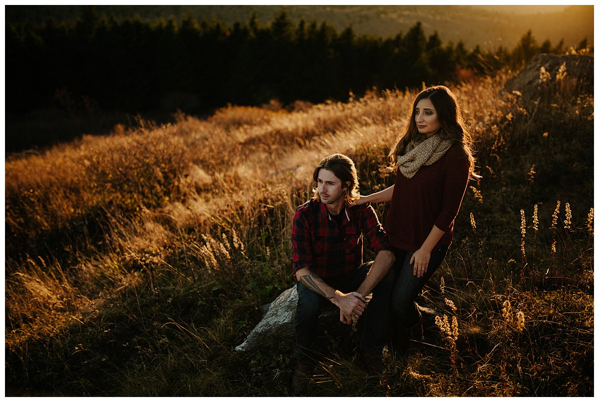 An engaged couple standing on a rock at sunset, both wearing burgundy with golden grass being hit by the sun.  They are currently planning their wedding during the coronavirus outbreak.   