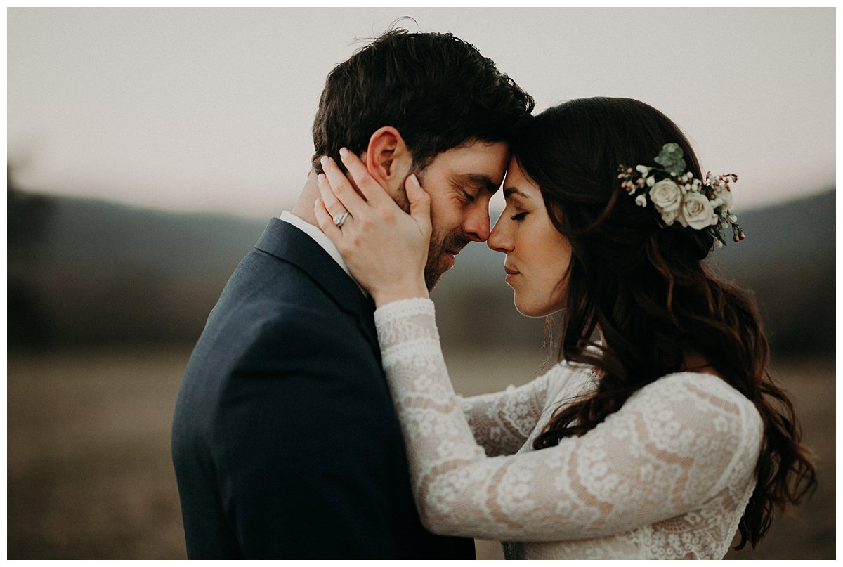 A bride and groom are touching foreheads and closing their eyes as they embrace one another.  The bride has a beautiful flower hair bow and a long sleeve wedding dress.  She is grabbing her groom's face.  