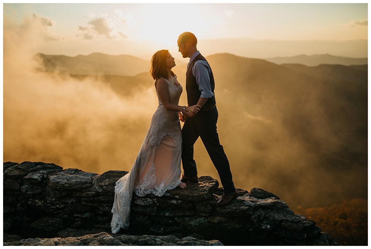 An elopement at Craggy Gardens at sunset in which a bride and groom are facing each other on a rock wall at sunset with the fog in the background.  An elopement is a great alternative when planning your wedding during coronavirus.   
