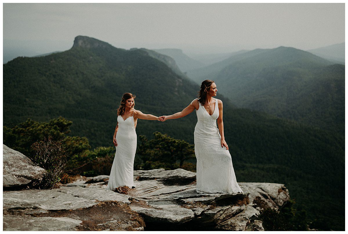 Two brides eloping at Hawksbill overlooking the beautiful mountains while holding hands.
