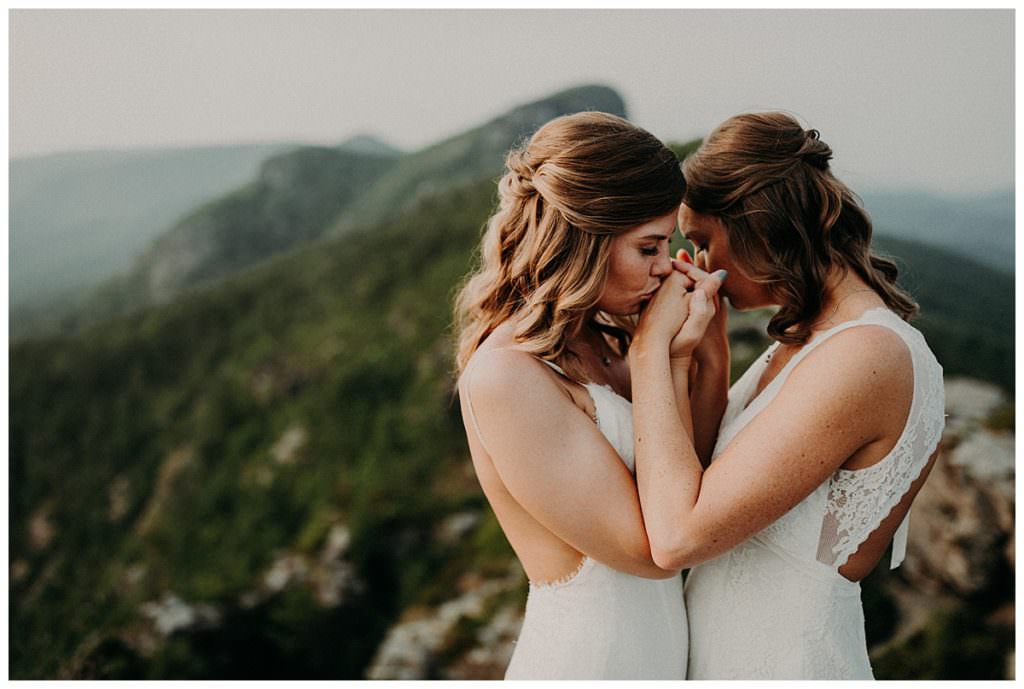 A closeup of Two brides holding both sets of hands and kissing each others hands atop a cliffside. 