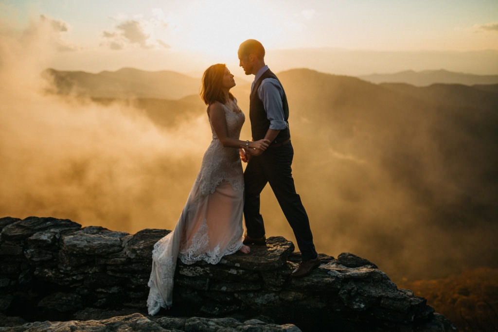 An Asheville elopement photographer captured a stunning image of a bride and groom at sunset with fog in the background. The Blue Ridge Mountains can be seen in the background.