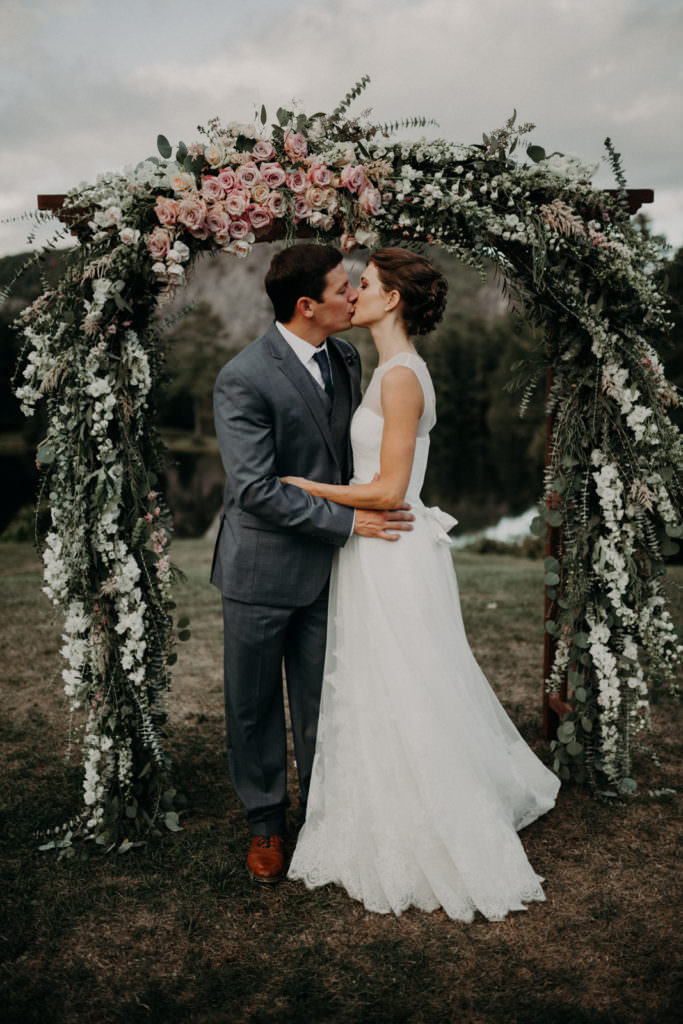 An Asheville elopement photographer captured an image of a couple kissing in front of a massive flower arbor. The image links to the photographer's wedding portfolio.