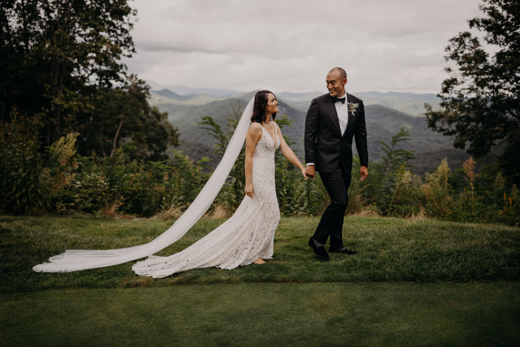 An Asheville wedding photographer captured a beautiful image of a bride and groom walking with the mountains in the background. The purpose of the image is to share a review of his Asheville wedding photography services.